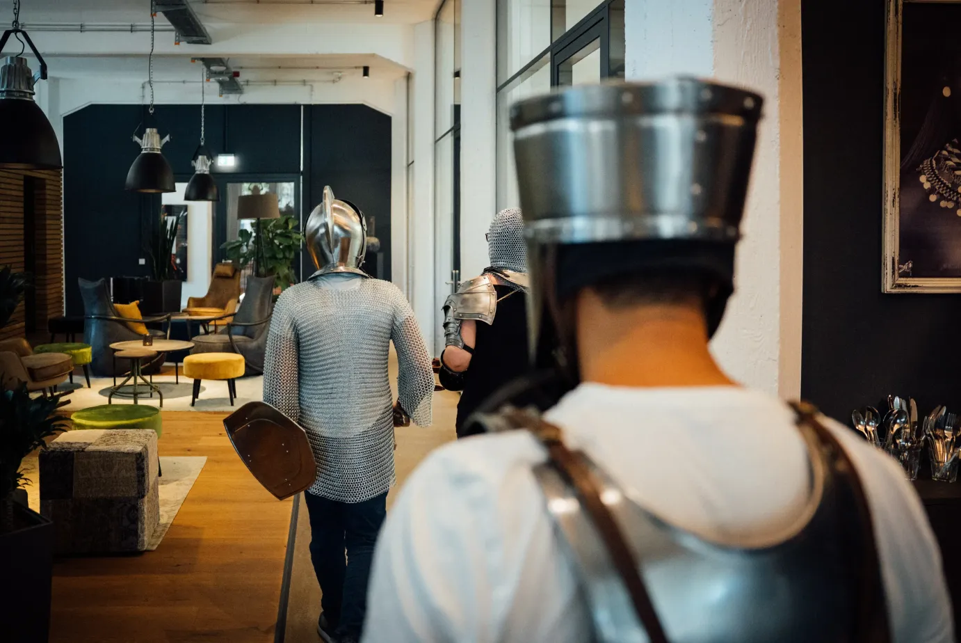 Three team members wearing medieval knight armor walk through a modern office, carrying shields and wearing chainmail and helmets